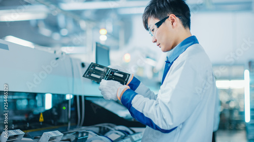 High-Tech Factory: Quality Control Engineer Checks Electronic Printed Circuit Board it for Damages. In the Background Assembly Line for PCB with Surface Mount Pick and Place Technology.