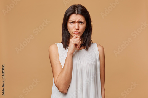 Indoor photo of serious suspicious tanned woman standing hand keeps chin, frowning, expressing distrustfulness and concern, over beige background