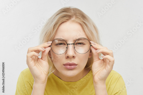 Portrait of a beautiful young woman with spectacles, looking curiously at you, squinting, trying to look closer. Girl with poor eyesight in yellow T-shirt posing on a white background in the studio