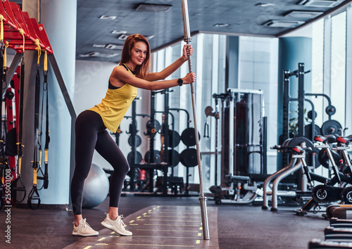 Beautiful slim fitness girl smiling and looking at a camera, posing for a camera while leaning on a barbell in the modern gym
