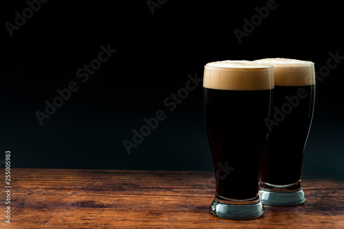 Alcohol abuse, bitter refreshing alcoholic beverage and dry stout concept theme with frothy glass pints of dark beer on wood table isolated on black background with copyspace in bar or pub setting