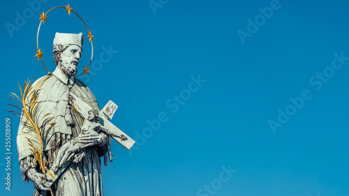 Statue of saint with cross at the Charles Bridge in Prague at blue sky, Czech Republic, summer time