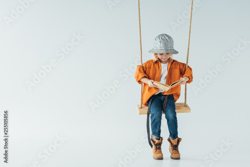 cute kid in jeans and orange shirt sitting on swing and reading book on grey background