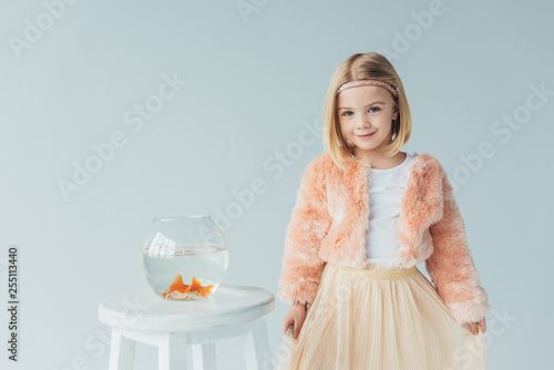 adorable kid in faux fur coat and skirt looking at camera and standing near to stool with fishbowl isolated on grey