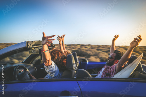 Side view group three euphoric friends in convertible car twisting and waving Two curly girls and charming bearded man on vacation having fun in topless auto Arms up palms open driving in the sunlight