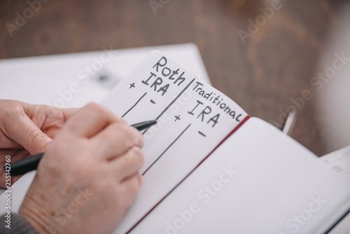 cropped view of senior woman writing in notebook with roth ira and traditional ira words