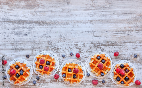 Traditional waffles with fresh raspberries and blueberries on lace doily on wooden background. Top view, copy space, flat lay .