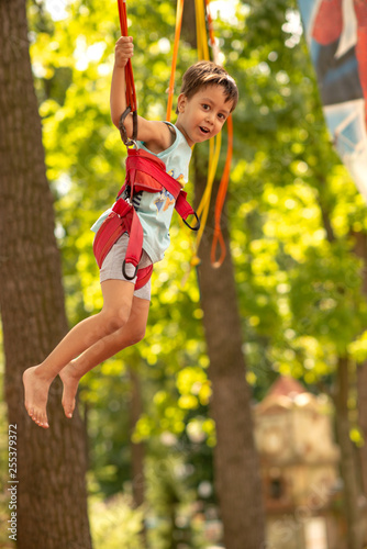 Emotional portrait of the boy with seat belts on an attraction "Trampoline". He with delight soars in air against the background of the green trees
