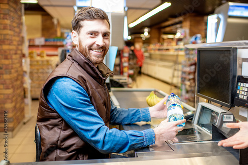 Portrait of a happy and cheerful man as a cashier, sitting at the cash register in the supermarket