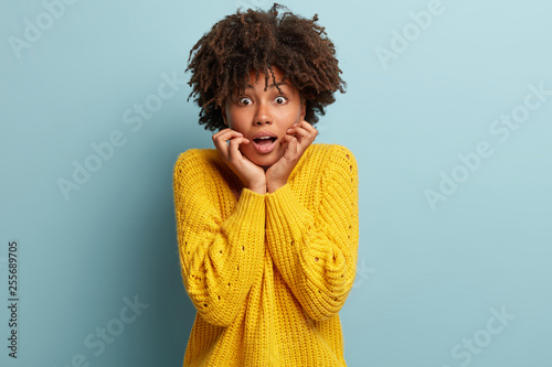 Wondered dark skinned lady puzzled and scared to make terrible mistake, holds both hands under chin, wears knitted warm sweater, isolated over blue wall, finds out awful truth, stares worried