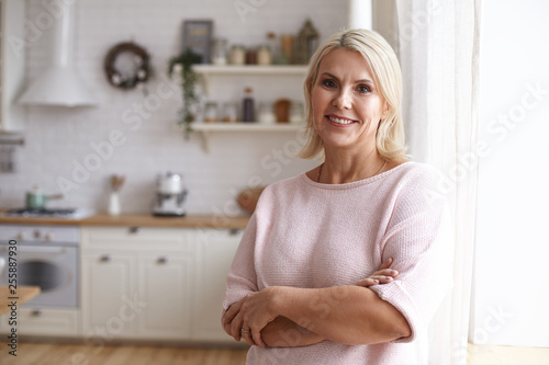 Attractive middle aged housewife with blonde hair and brown eyes posing indoors in her modern clean stylish kitchen interior, crossing arms on her chest and looking at camera with pleased smile
