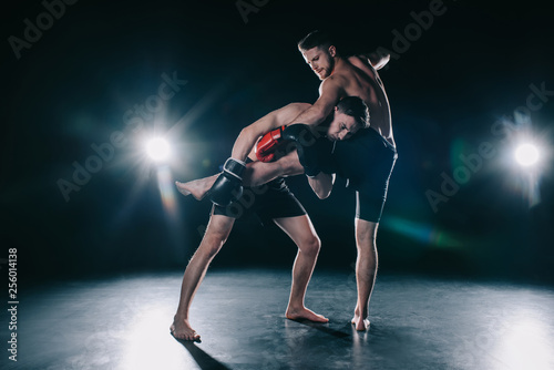 barefoot strong muscular mma fighter in boxing gloves clinching another while sportsman kicking him