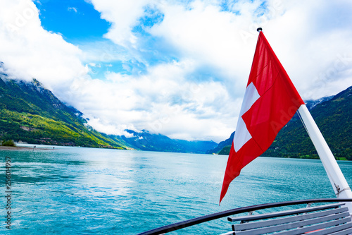 Red Swiss flag over landscape of lake Brienz in Interlaken, Switzerland from cruise in cloudy blue sky day