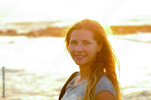 Candid portrait of young woman by the sea at sunset, strong sun backlight, background overexposed intentionally for atmosphere