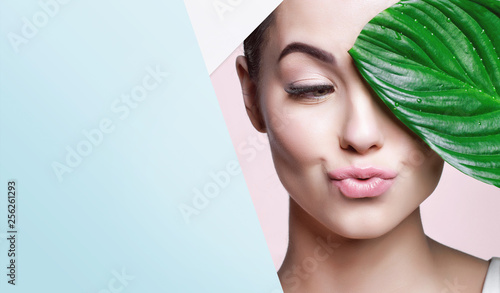 Portrait of young beautiful woman with healthy glow perfect smooth skin holds green tropical leaf, look into the hole of colored paper. Model with natural nude make up. Fashion, beauty, skincare.
