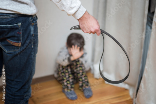 Family violence and aggression concept - furious angry man uses a belt for punishment scared or terrified child boy sitting at wall corner