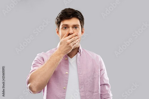 emotion, expression and people concept - shocked and speechless young man covering his mouth by hand over grey background