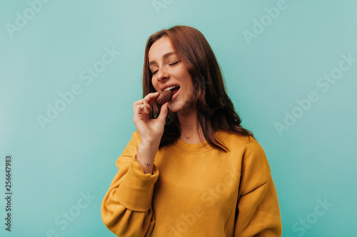 Indoor portrait of European woman with dark hair wearing bright clothes bitting a chocolate over blue background