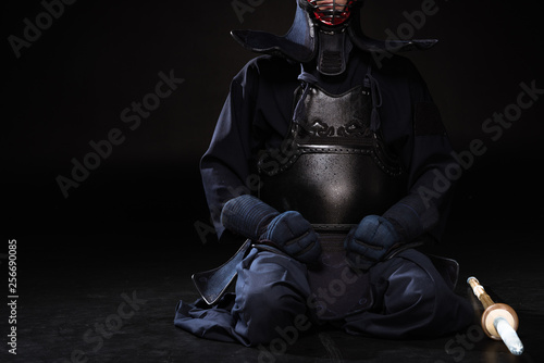 Partial view of kendo fighter in helmet with bamboo sword sitting on black