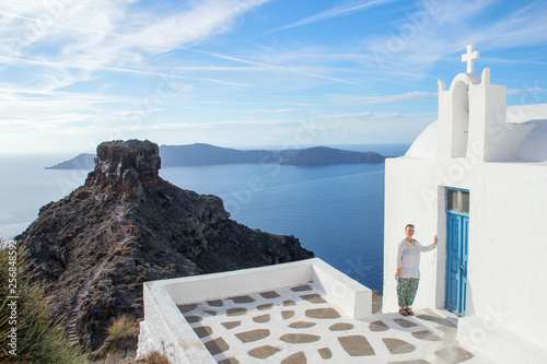 Santorini Island, Greece - November 17, 2016: A young girl tourist in white clothes is smiling next to a white church on the island of Santorini. Aegean Sea and Volcano on a summer day