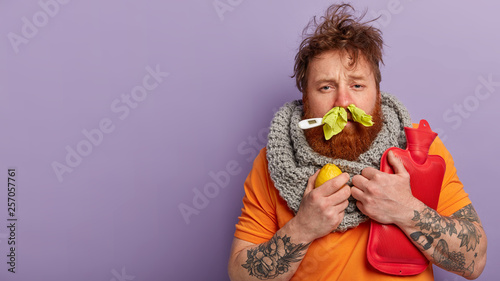 Photo of sick man has terrible sneezing, keeps napkins in nose, suffers from rhinitis, holds lemon and hot water bottle, keeps thermometer in mouth, has seasonal influenza virus, isolated on lilac