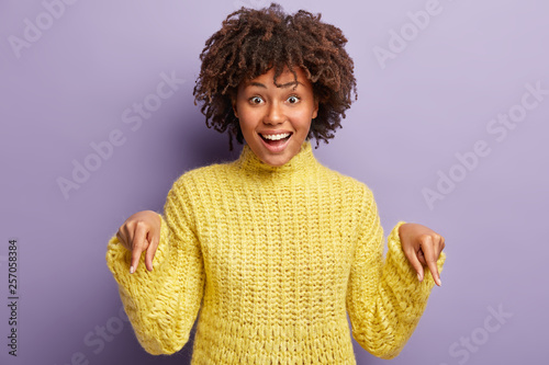 Satisfied dark skinned woman with curly hair, toothy smile, being in high spirit, points down on floor, likes something, expresses positive emotions, isolated over purple background. Promotion