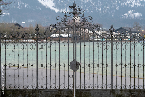 schönes, verziertes, schmiedeeisernes Tor vor dem Wolfgangsee, im Hintergrund Strobl und Blick auf die schneebedeckten Berge