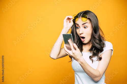 Oh, my! Gorgeous girl with a phone in her hand is surprised by something she saw on the screen is holding her glasses in front of bright yellow background.