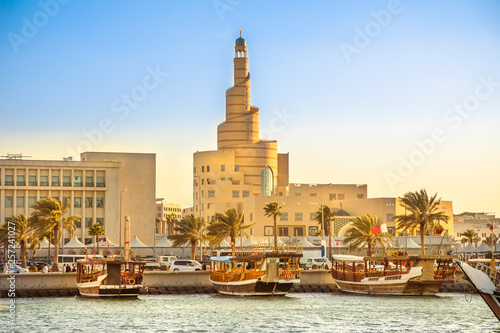 Traditional wooden dhow anchored at Dhow Harbor in Doha Bay with spiral mosque and minaret in the background at sunset. View from Corniche promenade. Qatar, Middle East, Arabian Gulf.