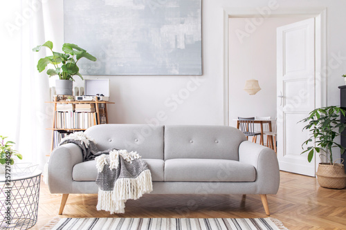 Modern scandianvian living room with design sofa with elegant blanket, coffee table and bookstand on the white wall. Brown wooden parquet. Concept of minimalistic interior. View on dining room. 