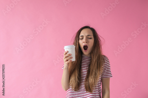 Cute girl with a glass of coffee in hand is yawning. She can't wake up.