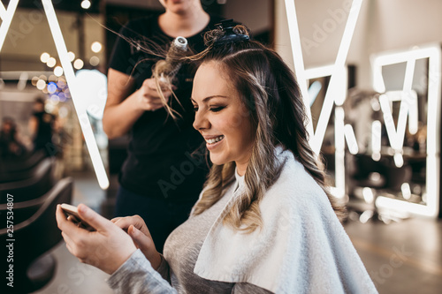 Beautiful woman with long hair at the beauty salon using smart phone and choosing hairstyle while getting a hair blowing. Hair salon styling concept.