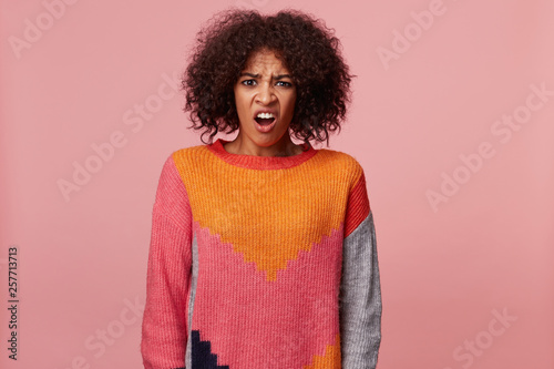 Emotional african american woman with afro hairstyle looking at something terrible awful dreadful disgusting, frowns her face, wearing colorful sweater, isolated over pink background