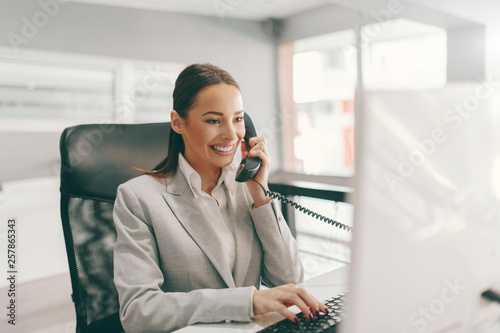 Beautiful Caucasian secretary in formal wear talking on the phone and typing on the PC while sitting in office. Success is not what you have, it's what you running for.