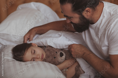 Loving father tucking blanket for his lovely little daughter. Adorable girl sleeping in bed, hugging her teddy bear, her father pulling up her blanket. Single parenting concept