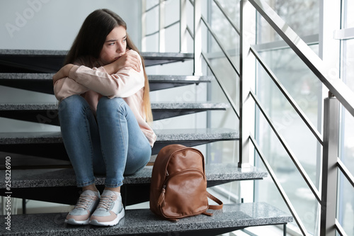 Upset teenage girl with backpack sitting on stairs indoors. Space for text