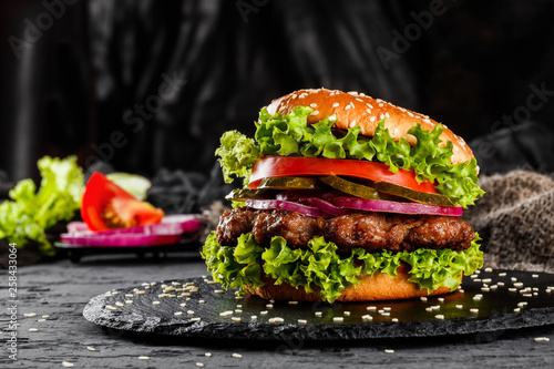 Beef burger with tomatoes, red onions, cucumber and lettuce on black slate over dark background. Unhealthy food