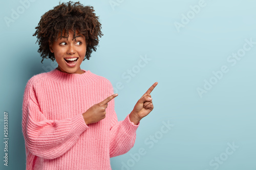 Horizontal shot of positive lovely woman with Afro haircut, points away with both fore fingers, demonstrates copy space for your information, has appealing look, sincere smile on face. Advertisement