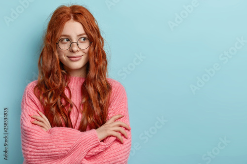 Pensive dreamy lovely young ginger female with pleasant appearance, imagines dreams come true, keeps hands crossed, dressed in oversized jumper, wears optical glasses, isolated over blue wall.