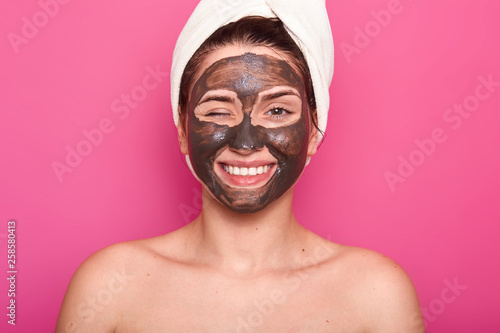 Close up portrait of beautiful young caucasian woman resting with chocholate anti acnee mask in wellnes spa salon, poses half nude, shows bare shoulders, winks with eye. Beauty and skin care concept.