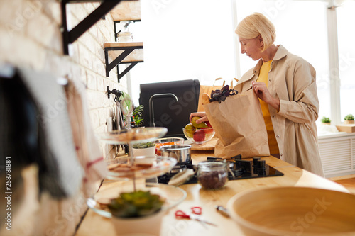 Content beautiful mature woman with short blond hair standing at kitchen counter and putting fruits in bowl while unpacking grocery bag