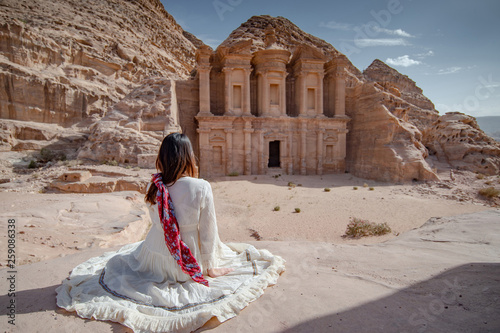 Asian woman tourist in white dress sitting and looking at Ad Deir or El Deir, the monument carved out of rock in the ancient city of Petra, Jordan. Travel UNESCO World Heritage Site in Middle East
