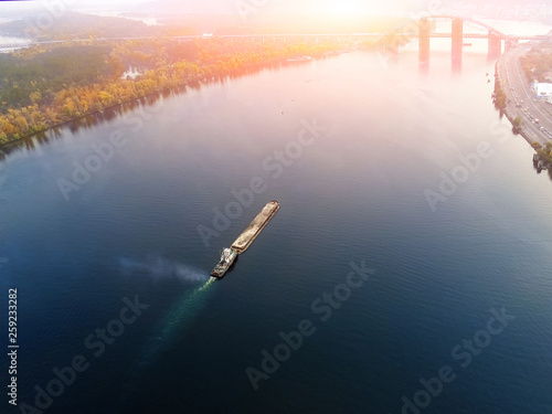 Scenic aerial cityscape of Kiev and river Dnipro at sunset. Tugboat supporting barge with sand bulk materials heading down river Dnieper. Ukrainian inland navigation and river freight cargo