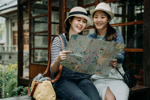 two female asian tourist with paper map sitting in bench by japanese style garden wooden house. Beautiful smiling girl friends traveler holding guide in hands discussing while travel kyoto japan