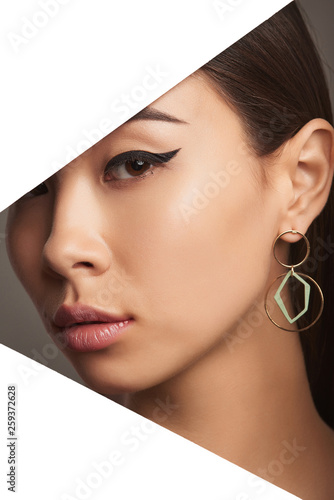Cropped closeup side portrait of Asian lady with black flicks, tilting her head behind triangle-shaped foreground. The pretty girl is wearing long dangle earring, adorned with 2 ring-shaped pendants.