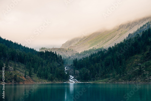 Ghostly forest near mountain lake in early morning. Mountain creek flows into lake. Ripple on smooth water surface. Low clouds. Dark calm atmospheric misty woodland landscape. Tranquil atmosphere.