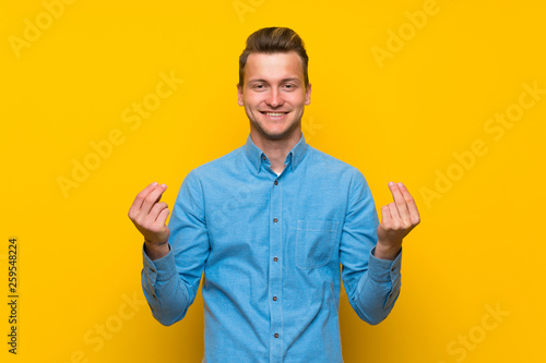 Blonde man over isolated yellow wall making money gesture