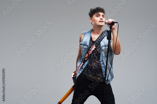 Portrait of rock singer man keeping electric guitar and static acoustic mic, sings a song loudly on grey background. Concept of rock music and rave