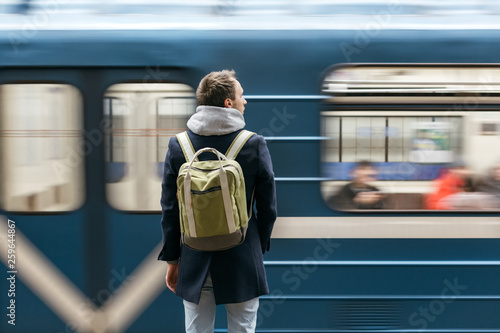 Lonely adult handsome man in a blue coat with a green backpack behind standing on metro platform, train in motion. Way to work for a modern city dweller. Morning commute. Urban life concept.