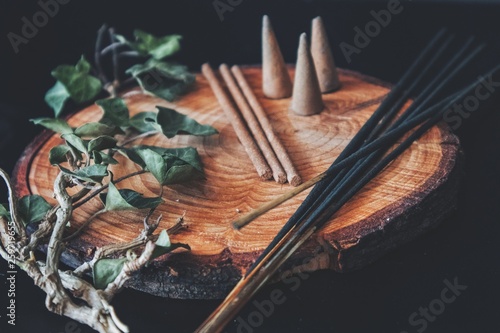 Examples of various types of incense - long black sticks, short hand made sticks and small triangular cones. Black background, placed on a wooden plate cutting with ivy plant branch laying next to it